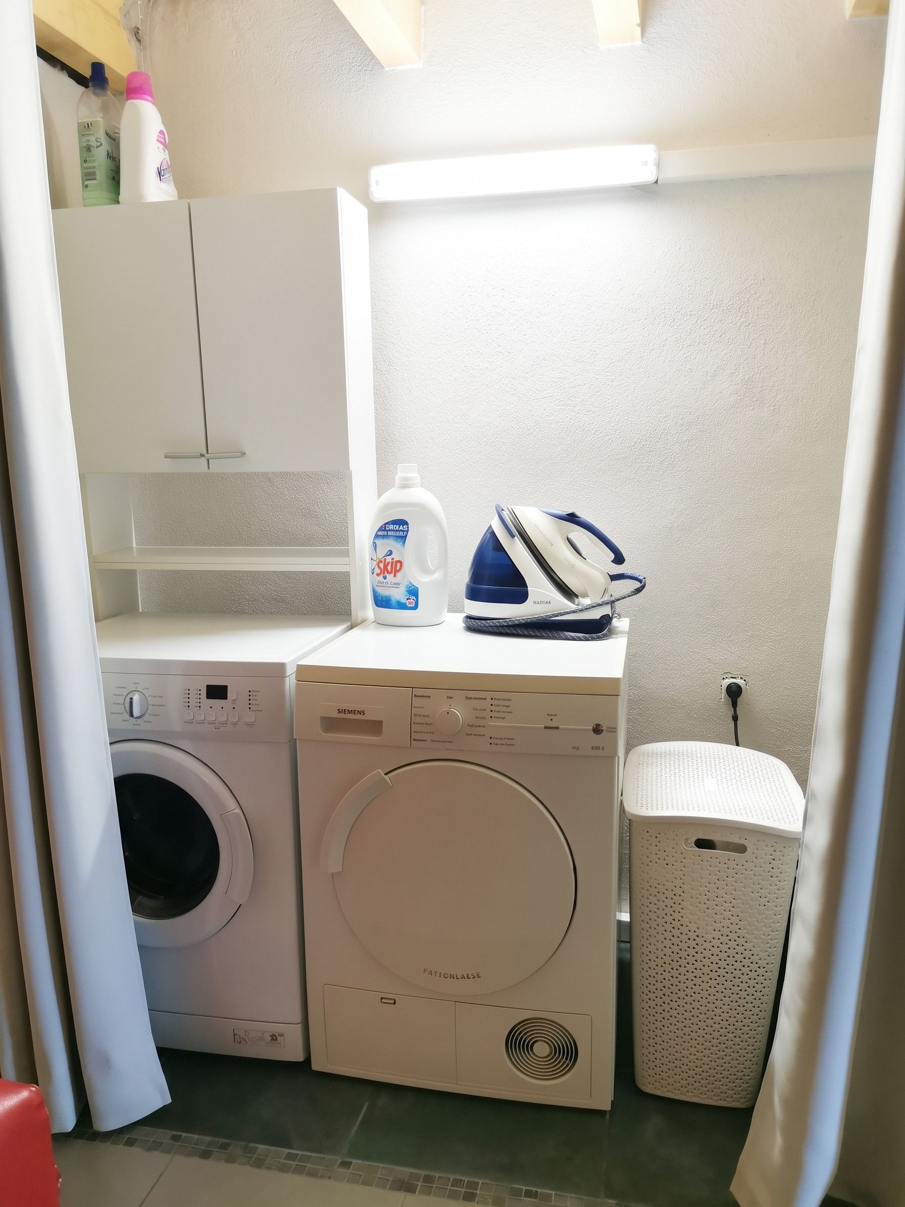 Een kleine wasruimte met een wasmachine, een strijkijzer en een fles wasmiddel.