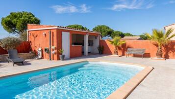 Une piscine à l’eau d’un bleu limpide, entourée d’une terrasse avec un fauteuil de relaxation et un petit bâtiment doté d’un toit en tuiles.