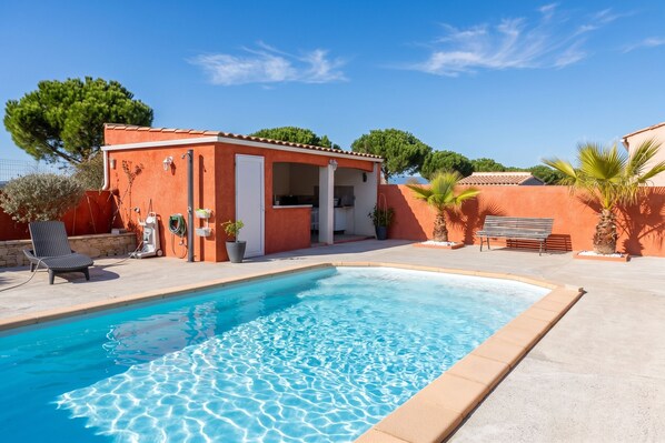 A swimming pool with a clear blue water surface, surrounded by a patio area with a lounge chair and a small building with a tiled roof. - Homerez - Villa in Carcassonne (Carcassonne)