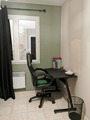 A room with a desk, chair, window, and a trash bin.
