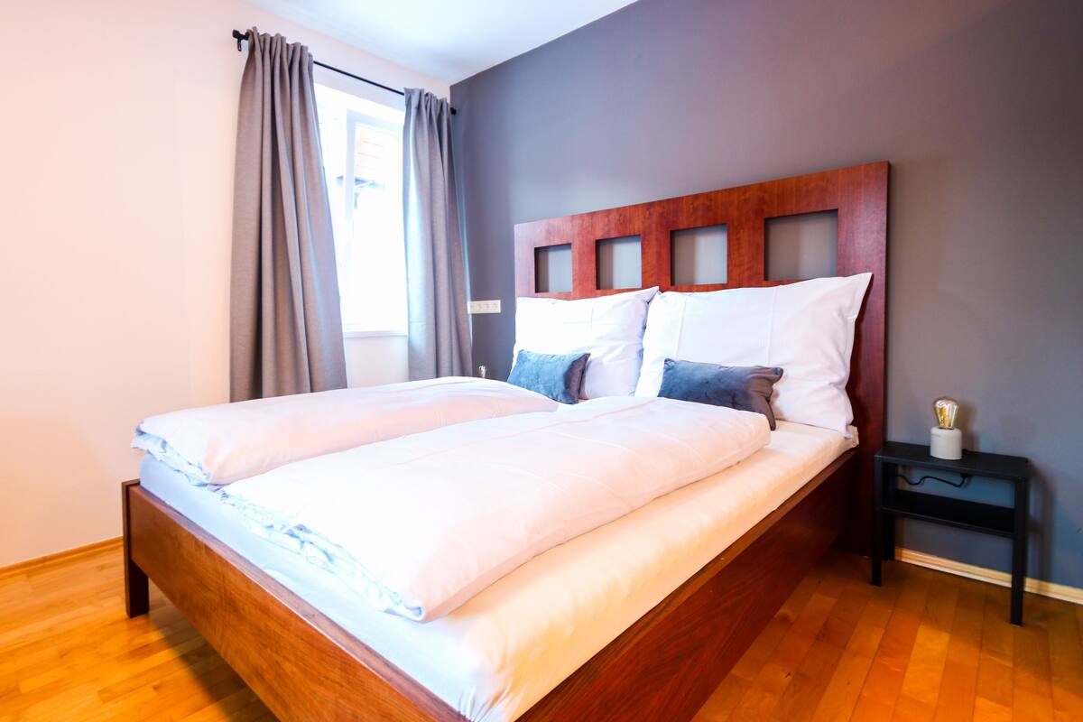 A double bed with a wooden headboard, grey curtains, and a bedside table with a lamp.