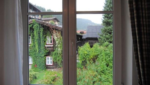 A window with a view of a traditional building with a wooden facade and a balcony, surrounded by greenery and trees. - Appartement Bosek (Wagrain)