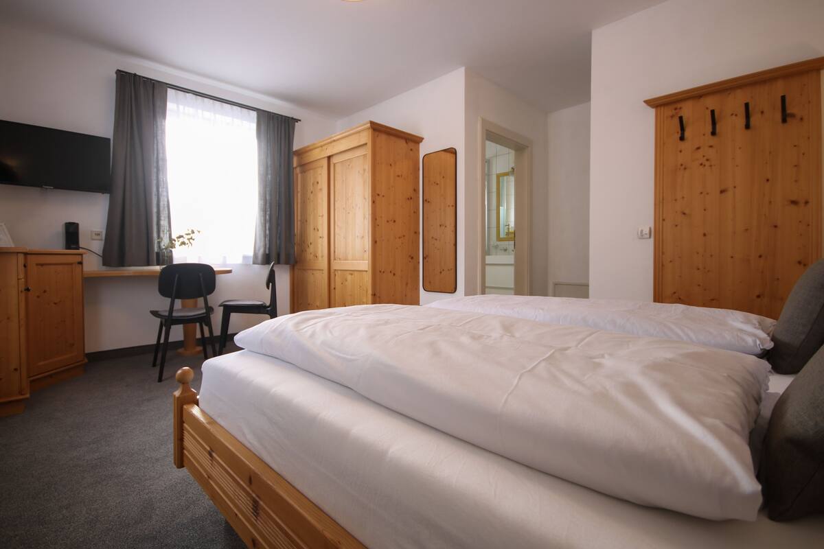 A bedroom with a large bed, wooden wardrobe, a desk with a chair, and a television on the wall.