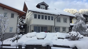 A two-story building with a snow-covered roof and windows, surrounded by snow-covered trees and a wooden fence. - Appartement Bosek (Wagrain)