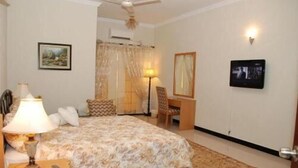 A hotel room with a bed, a desk, a chair, a lamp, a television, and a ceiling fan. - Savoy Inn (Islamabad)