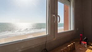A room with a large window overlooking a beach, a wooden table with a vase and a jar, and a door with a handle.