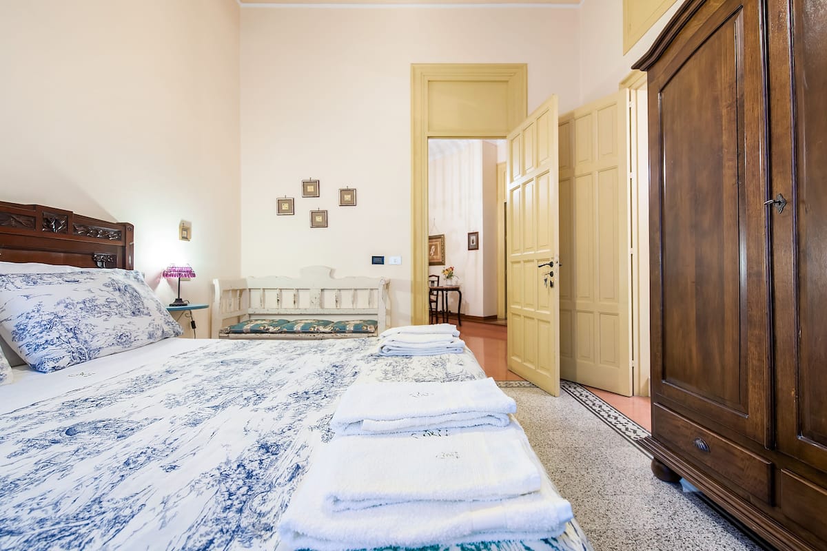 A bedroom with a bed, a bedside table, a lamp, a wardrobe, and a door leading to another room.