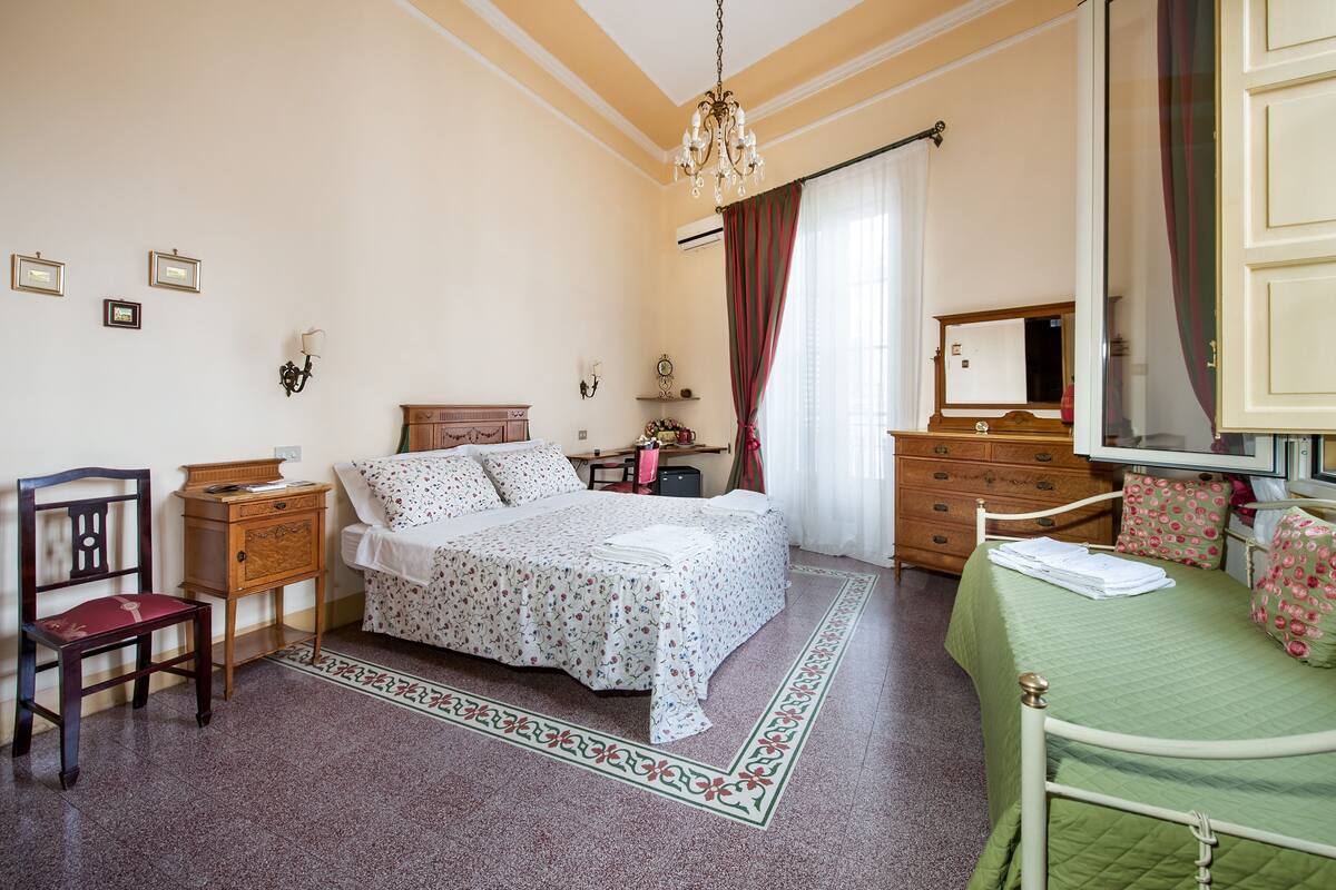 A bedroom with a bed, a chair, a dresser, a nightstand, a mirror, and a chandelier.
