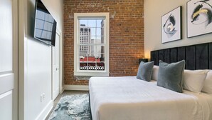 Un dormitorio con pared de ladrillo, ventana con vistas a edificios, cama con almohadas y televisor montado.