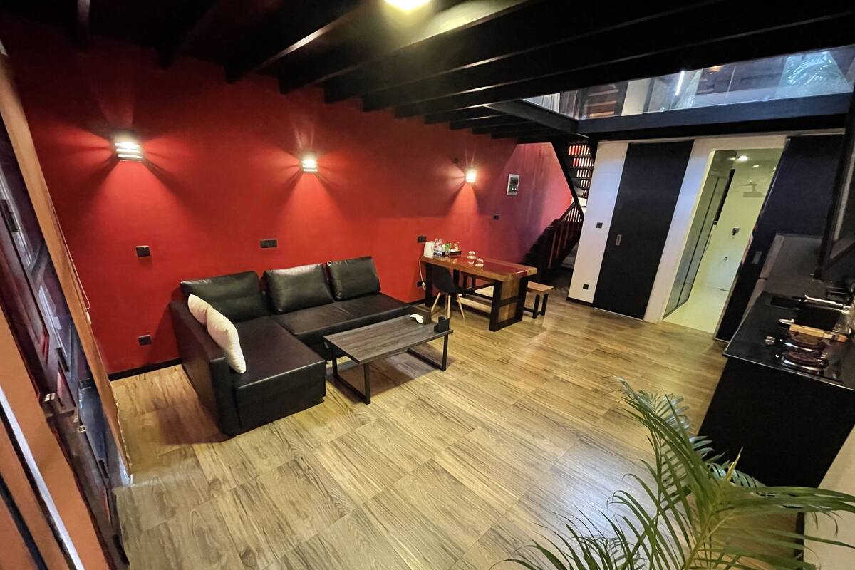A modern living room with a black leather sofa, wooden coffee table, and a dining table. There is a staircase leading to an upper level.