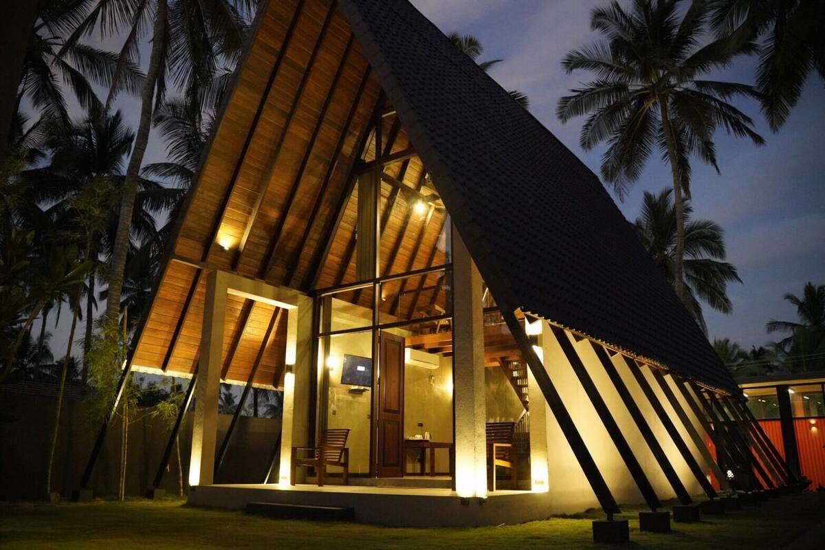 A modern, triangular-shaped building with large glass windows and wooden beams, surrounded by palm trees.