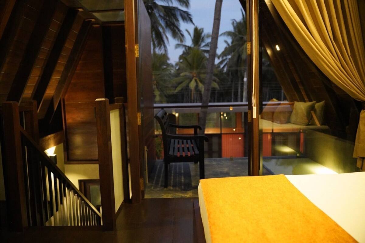 A room with a bed, a staircase, and a balcony with a view of palm trees.