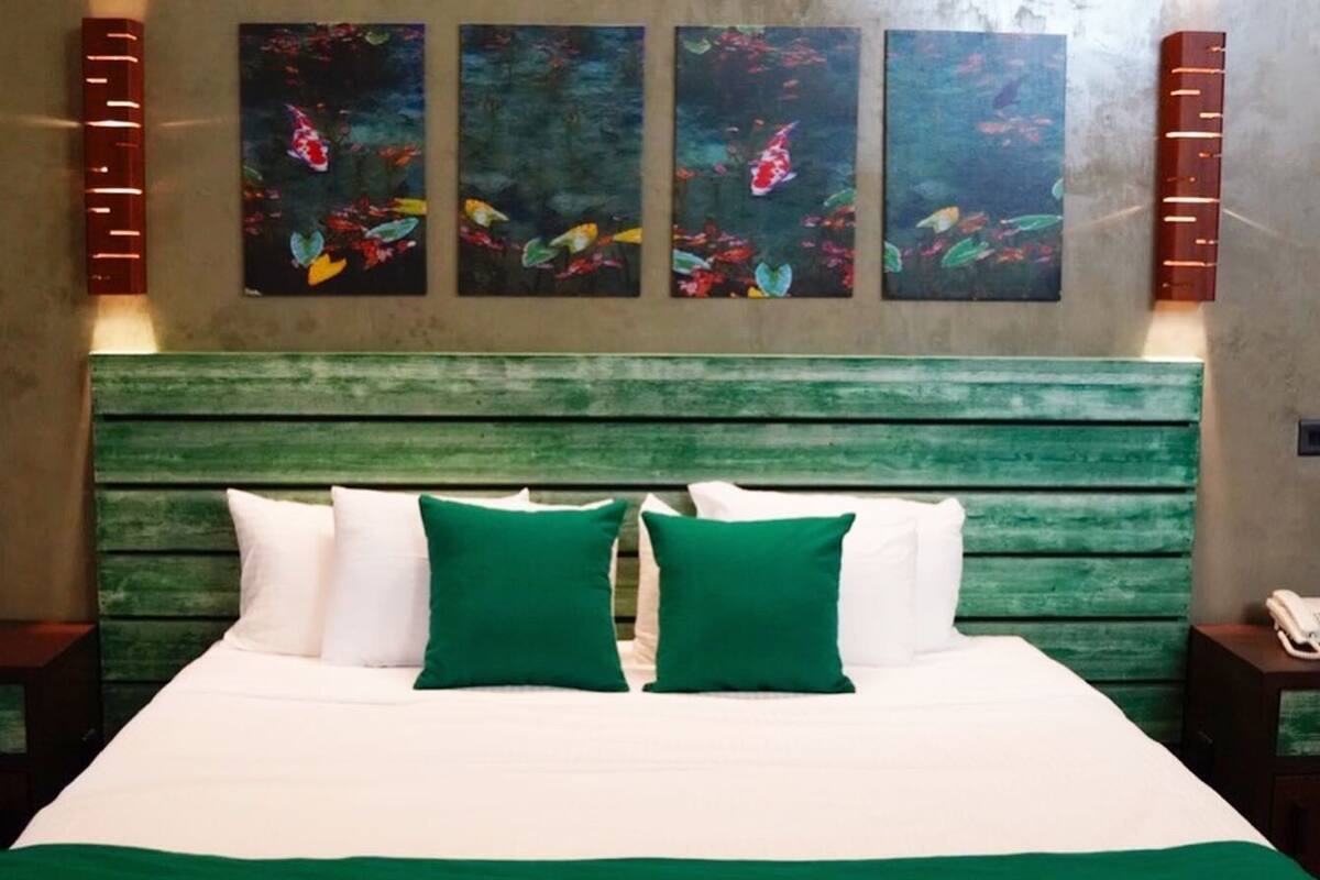 A hotel room with a bed, green headboard, white bedding, and three paintings on the wall.