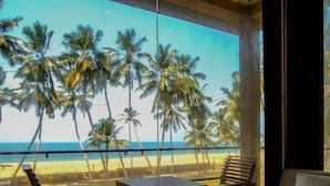 A room with a large window overlooking a beach with palm trees. There are two wooden tables and two chairs. - Soluna Beach Resort (Marawila)