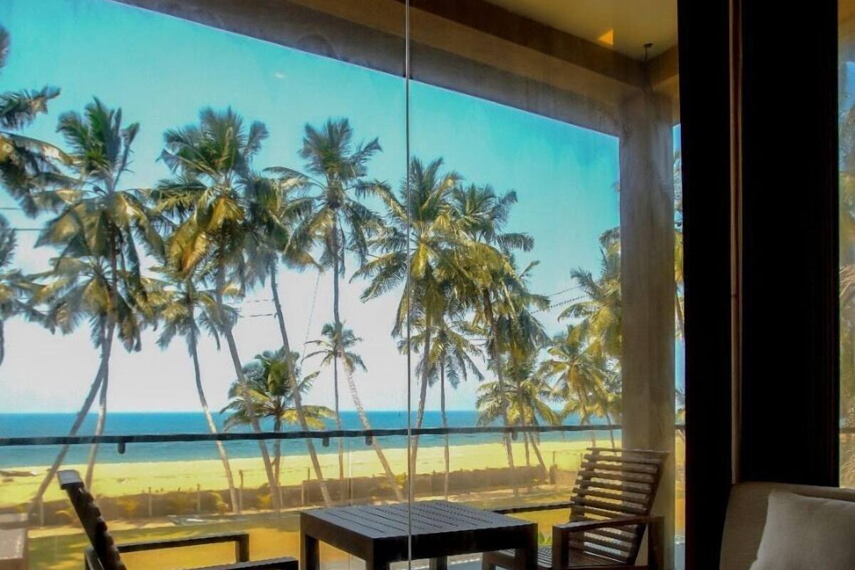 A room with a large window overlooking a beach with palm trees. There are two wooden tables and two chairs.