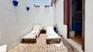A small outdoor area with two white lounge chairs, a small table with a bottle and a plant, and a blue bucket on the wall.