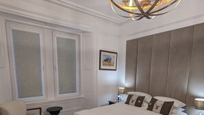 A bedroom with a large bed, a chair, a small table, a chandelier, and a window with frosted glass. - Cambeth Lodge Guesthouse (Inverness)