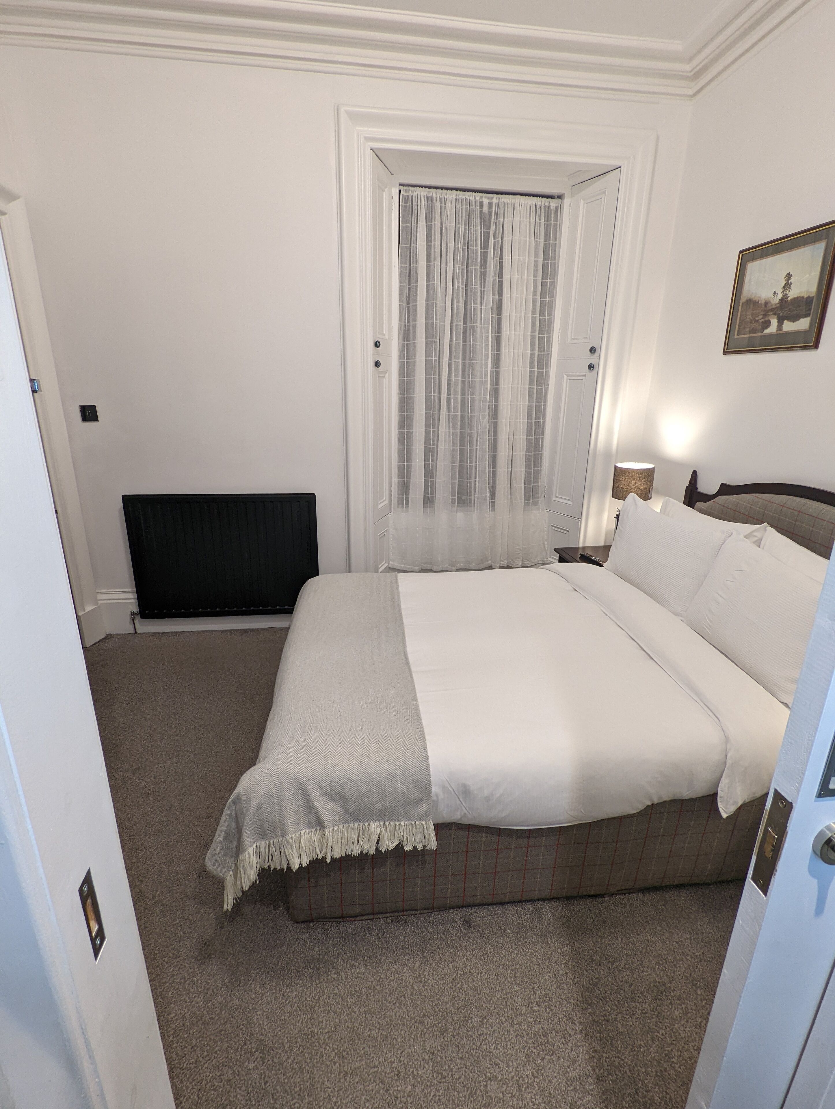 A bedroom with a bed, a radiator, a window with sheer curtains, a bedside lamp, and a framed picture on the wall.