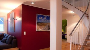 Un intérieur moderne avec un mur d’accent rouge, un escalier en bois et une vue sur la plage, visible à travers une fenêtre.