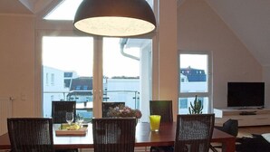 Un coin repas avec une table dressée pour deux personnes, deux chaises en osier, un grand luminaire suspendu et une vue sur les bâtiments à travers les fenêtres.