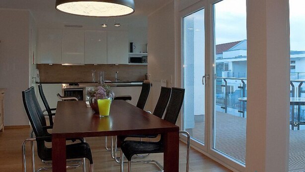 Un espace repas moderne avec une table et des chaises en bois, un grand luminaire suspendu et une vue sur le balcon à travers des portes vitrées.