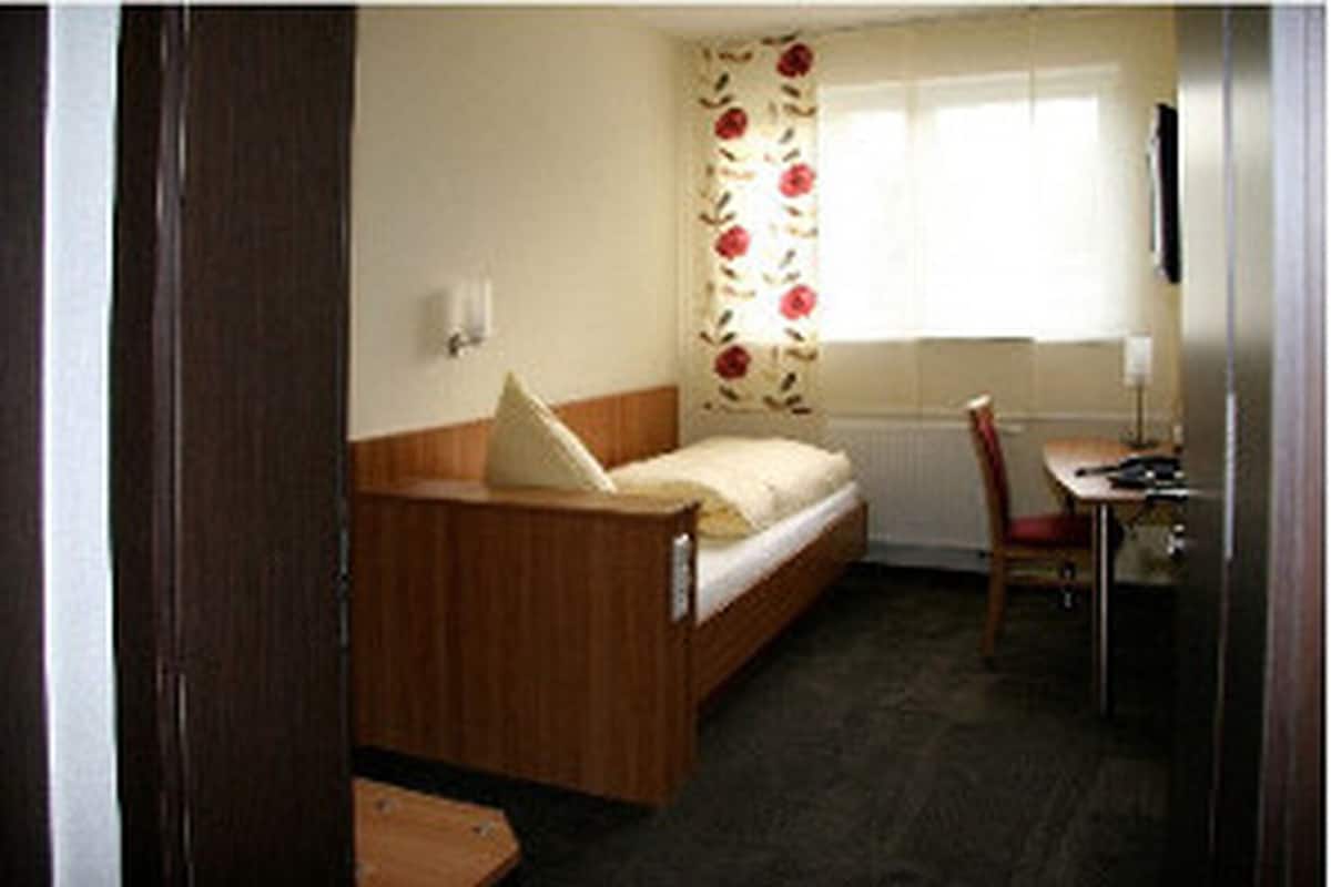 A small, single-bed room with a wooden bed, a desk with a chair, a radiator, and a window with floral curtains.
