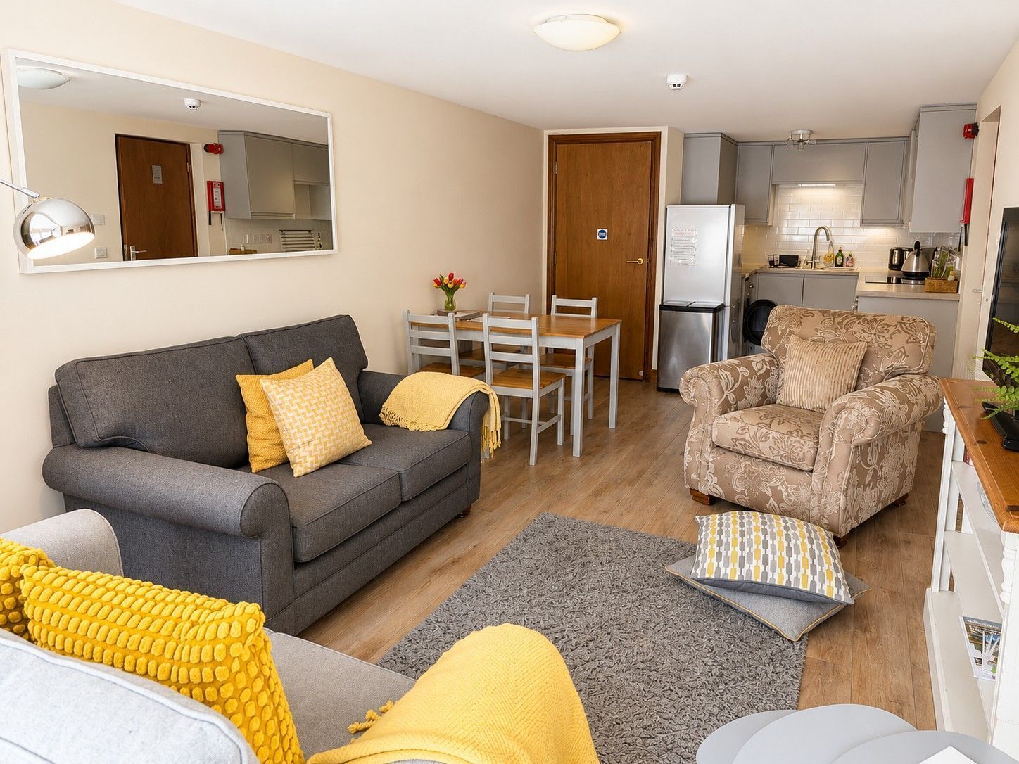 Ein Wohnzimmer mit grauem Sofa, einem gemusterten Sessel, einem Esstisch mit Stühlen, einer Küchenzeile mit Kühlschrank und einem Spiegel an der Wand.