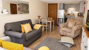 Ein Wohnzimmer mit grauem Sofa, einem gemusterten Sessel, einem Esstisch mit Stühlen, einer Küchenzeile mit Kühlschrank und einem Spiegel an der Wand.