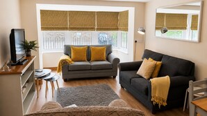 Un salon avec un canapé, un téléviseur, une petite table et une fenêtre avec des stores.