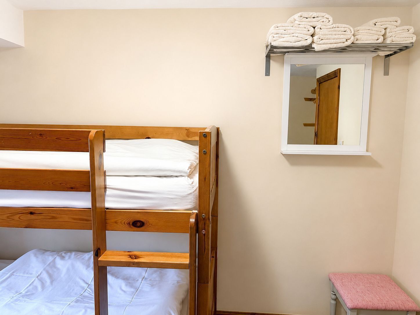 Een stapelbed met witte lakens, een klein plankje met opgevouwen handdoeken, een spiegel en een roze bankje.