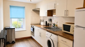 A compact kitchen with a washing machine, microwave, oven, and a window with blinds. - Howgills Apartment 9 (self-catering accommodation with free parking and Wi-Fi) (Sedbergh)
