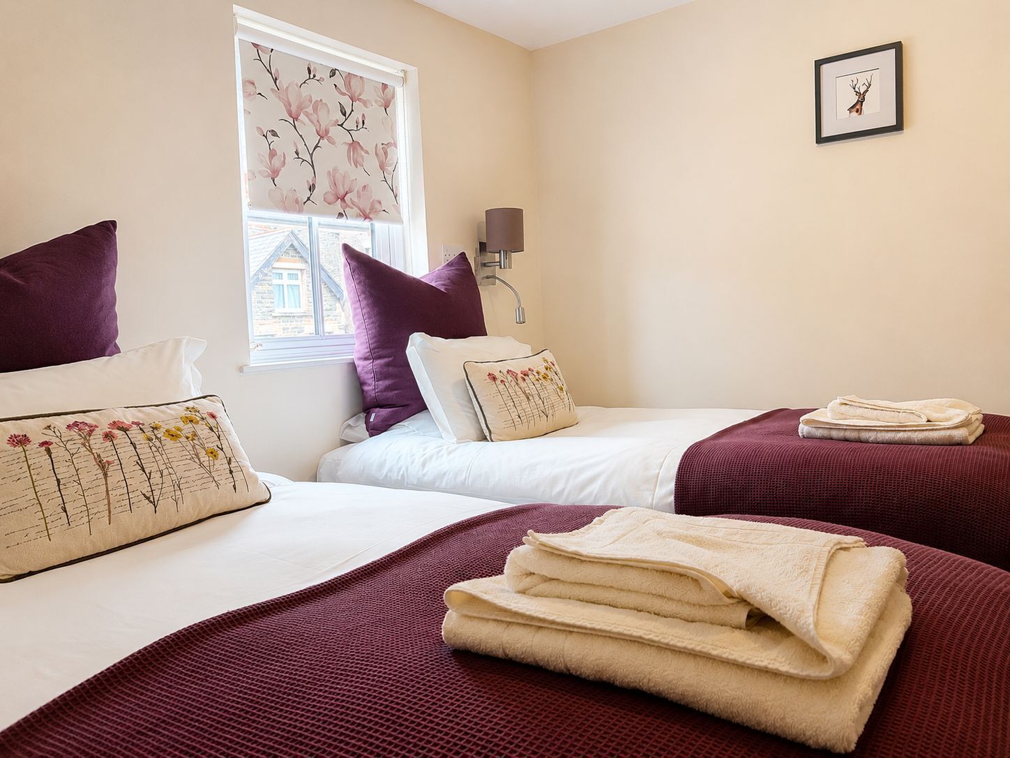 A hotel room with two beds, a window with floral blinds, a wall-mounted lamp, and a framed picture.