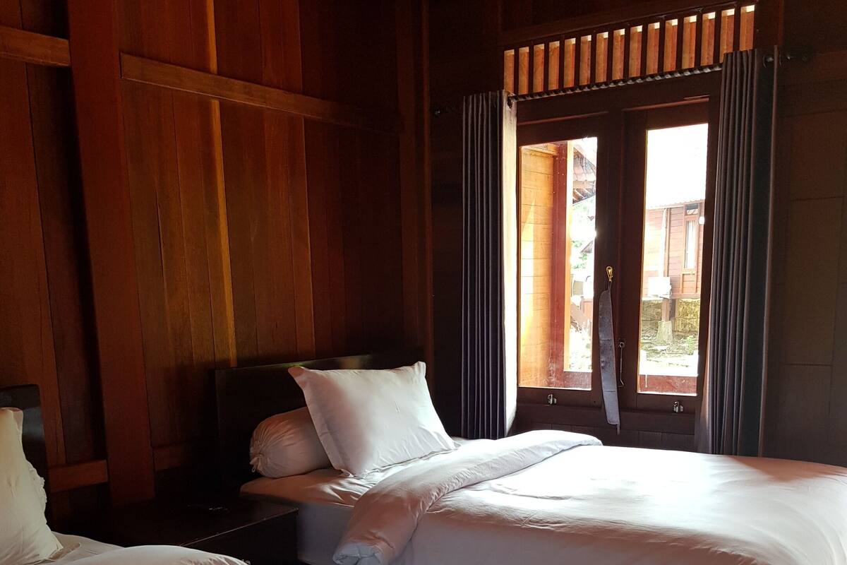 A room with two beds, wooden walls, a window with curtains, and a view of the outside.