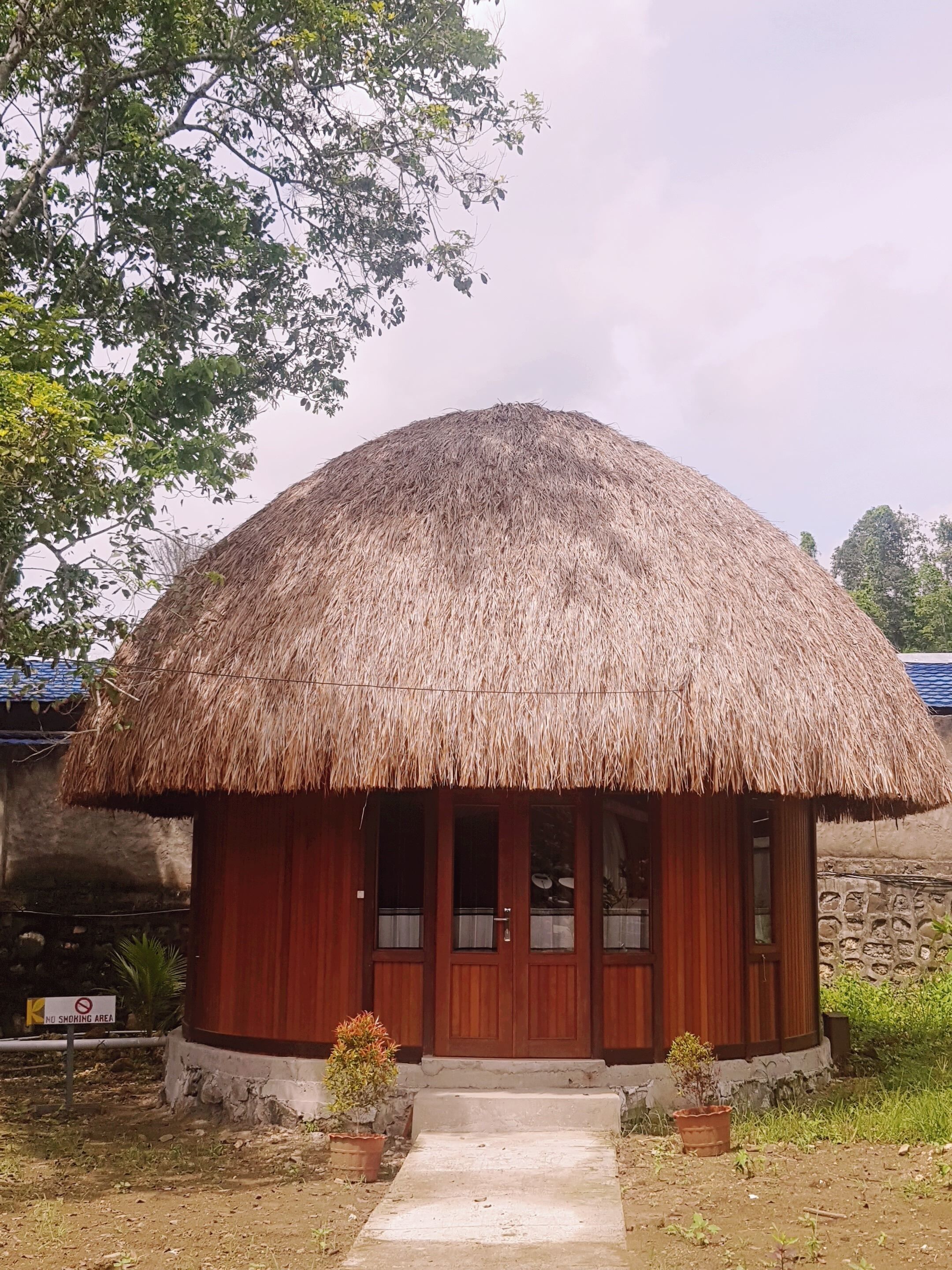 A thatched-roof hut with a wooden door and windows, surrounded by a garden and a stone wall.