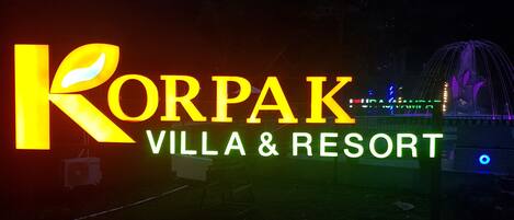 A sign for Korpak Villa & Resort with illuminated letters and a fountain in the background.