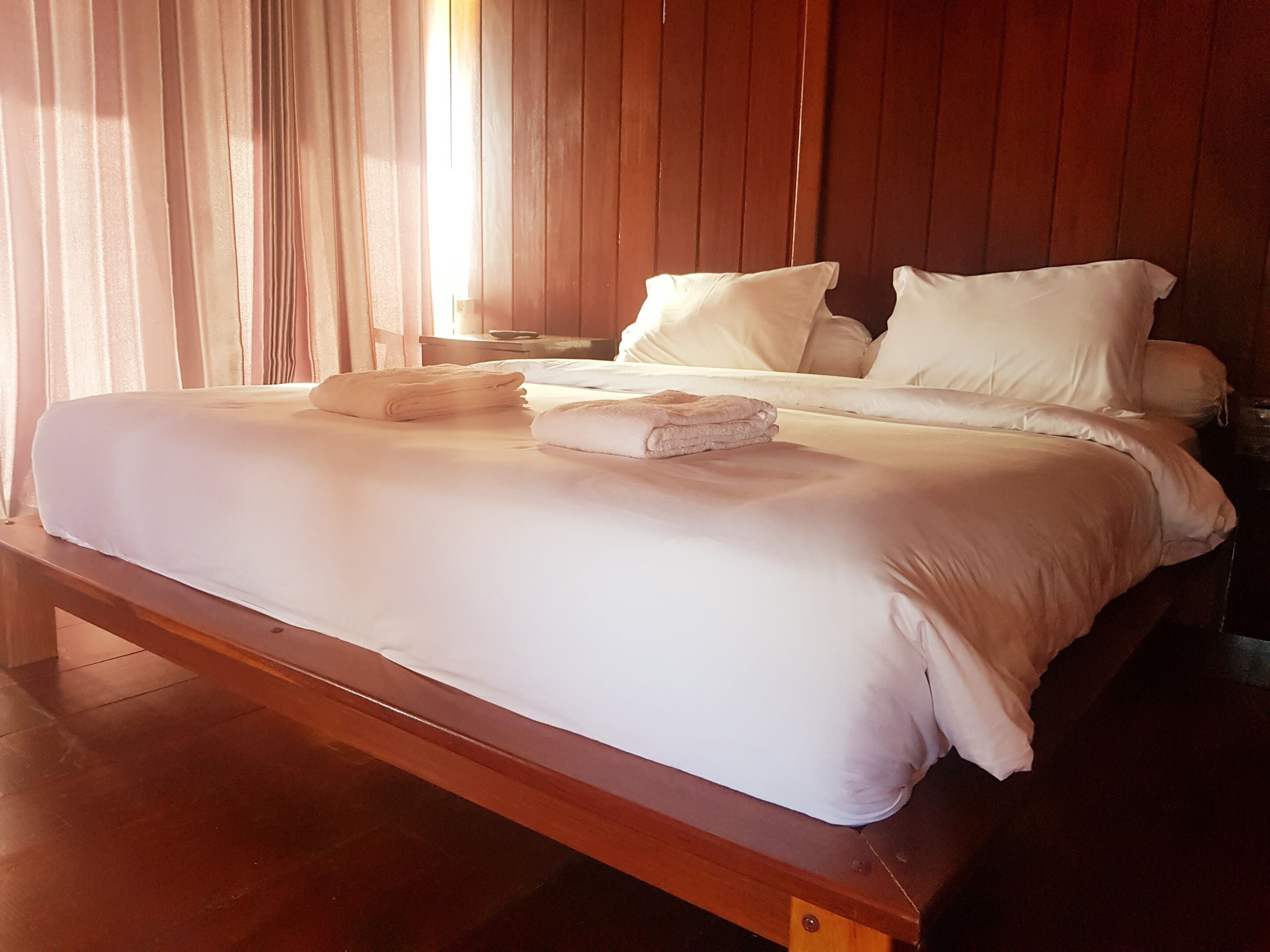 Una cama bien hecha con sábanas blancas y dos toallas dobladas, cabecera de madera y suelo de madera.