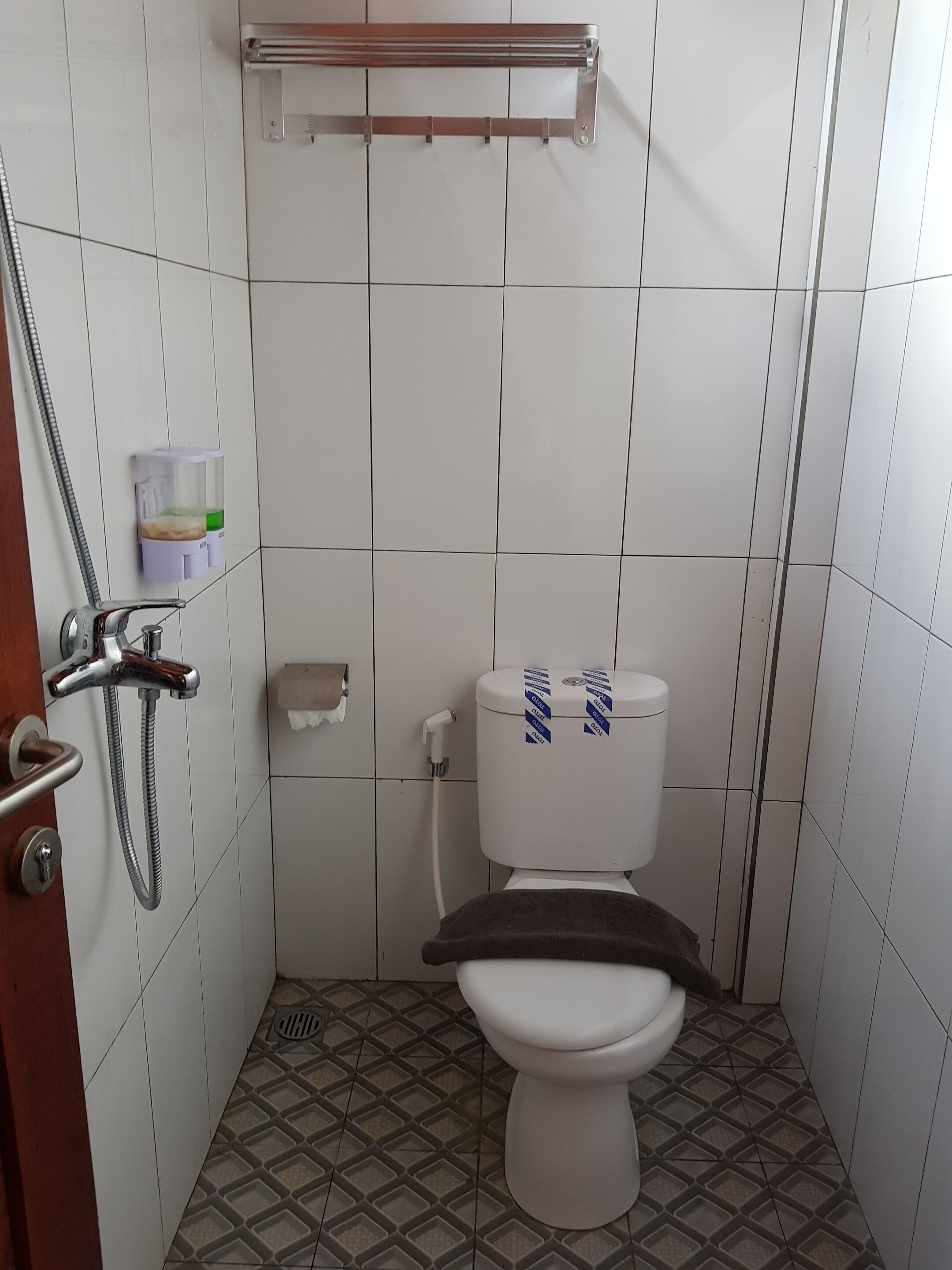 Un bagno piccolo con pareti rivestite di piastrelle bianche, un WC con coprisella marrone, una doccia con soffione manuale e un porta asciugamani sopra il gabinetto.