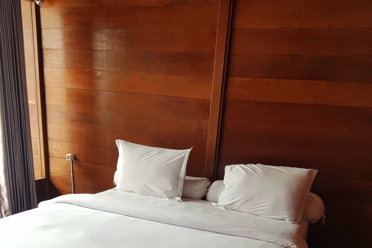 A large bed with white linens and multiple pillows, positioned against a wooden wall with a window on the left.