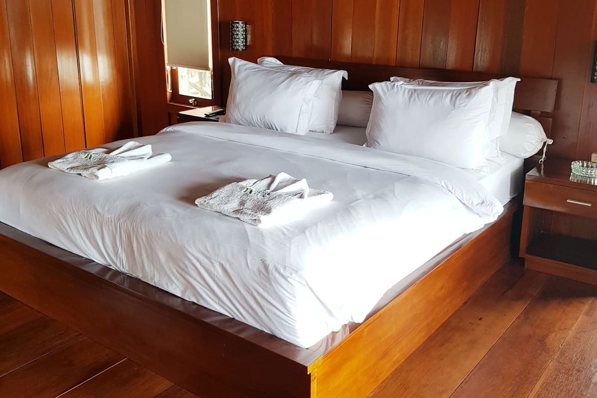 A large wooden bed with white linens and towels, a nightstand with a lamp, and a wooden floor.