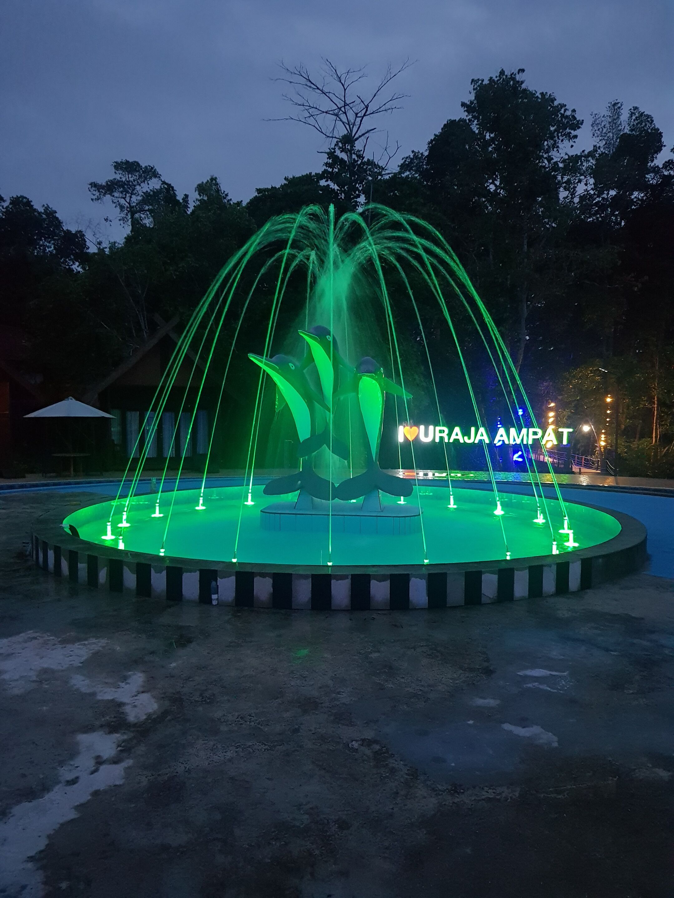 A fountain with a circular base and multiple water jets, surrounded by a paved area.