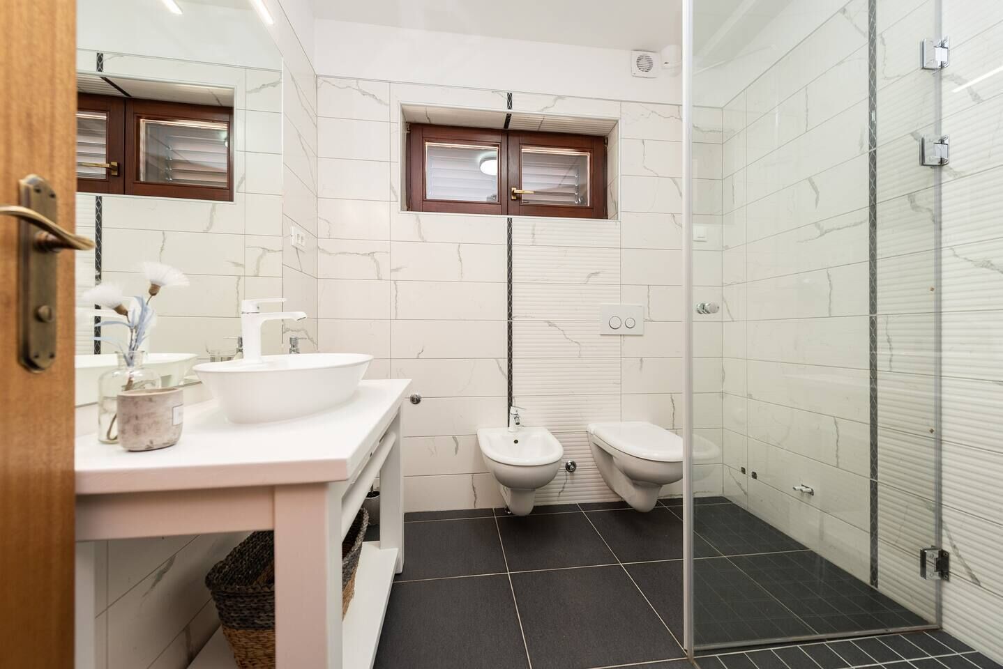 Un baño moderno con un lavamanos blanco, dos inodoros, una ducha y una ventana pequeña.