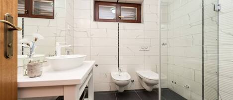 Un baño moderno con un lavamanos blanco, dos inodoros, una ducha y una ventana pequeña.