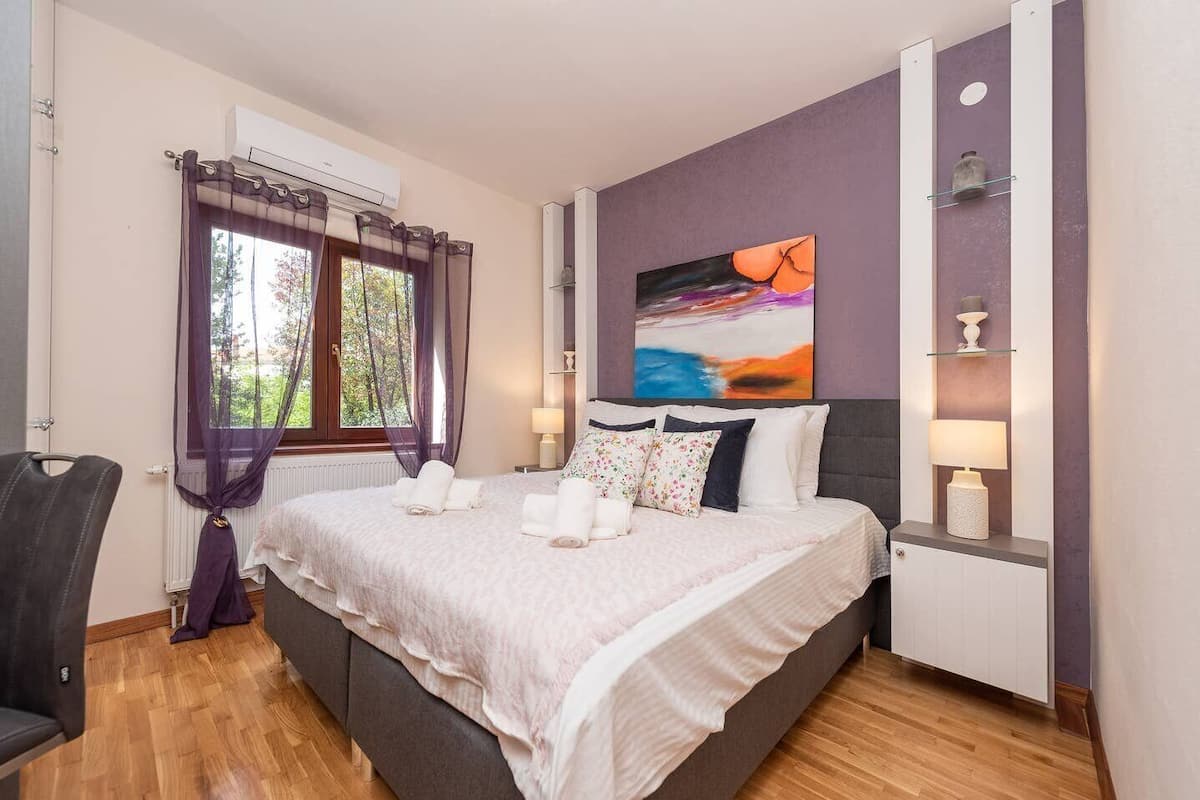 A bedroom with a bed, a chair, a window with curtains, a painting, and a shelf with a lamp.