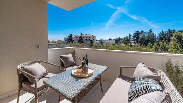 A balcony with wicker furniture, a table with a bottle and flowers, and a view of buildings and trees. - Zen Luxury Apartments (Zadar)