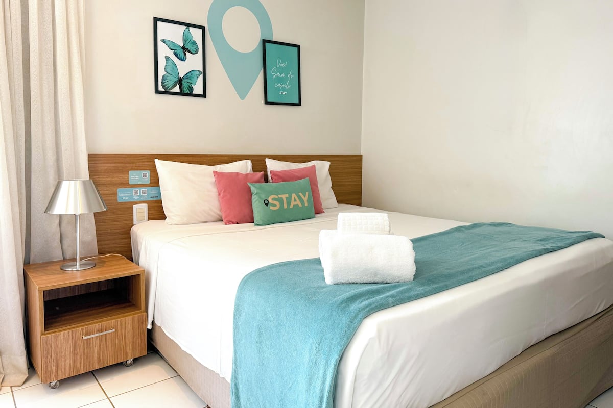 A neatly made bed with a teal throw blanket, a white towel, and a pillow with the word "STAY".