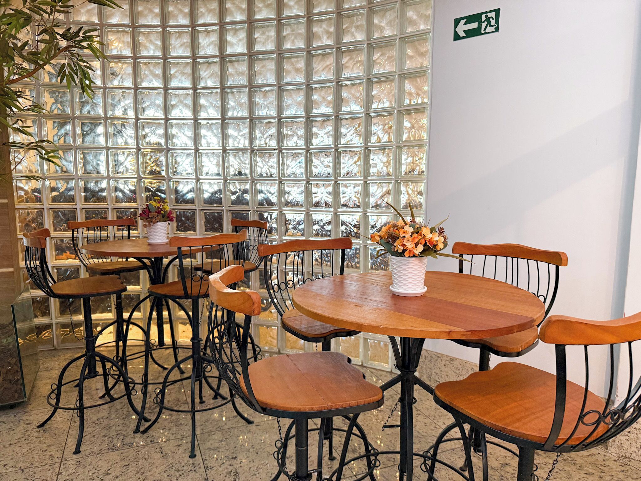 A restaurant with round wooden tables and metal chairs, a white vase with flowers on one table, and a large window with a grid pattern.