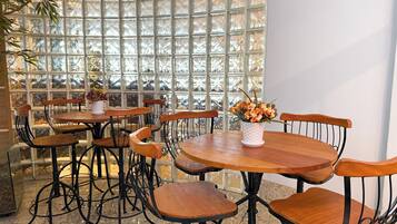 A restaurant with round wooden tables and metal chairs, a white vase with flowers on one table, and a large window with a grid pattern.