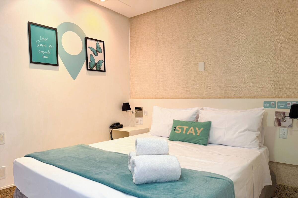 A hotel room with a bed, a bedside table with a lamp, a phone, and a wall with a 'STAY' sign.