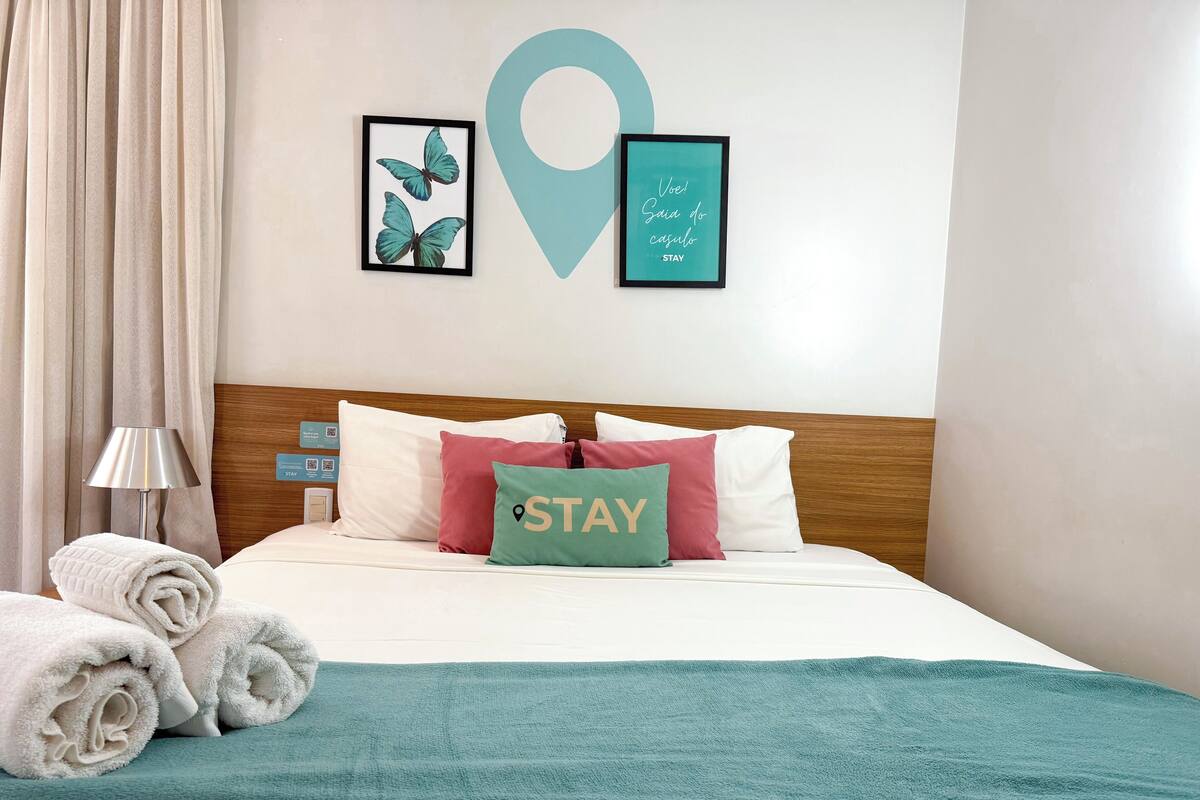 A neatly made bed with a teal throw blanket, pillows with a 'STAY' sign, and a framed butterfly artwork on the wall.