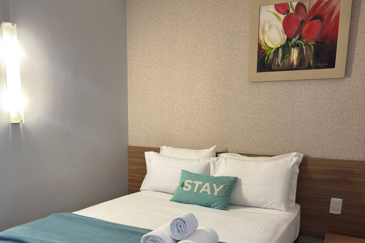 A neatly made bed with a green blanket, white pillows, and a 'STAY' pillow, a painting of flowers on the wall, and a bedside table with a lamp.